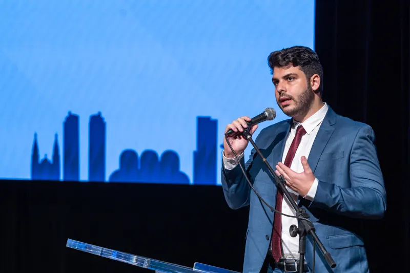 Allan Turano fala ao microfone em um palco, durante palestra