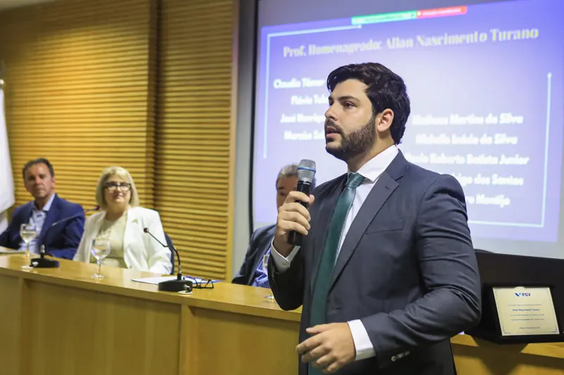 Allan Turano discursa ao microfone diante de slide que o apresenta como professor homenageado