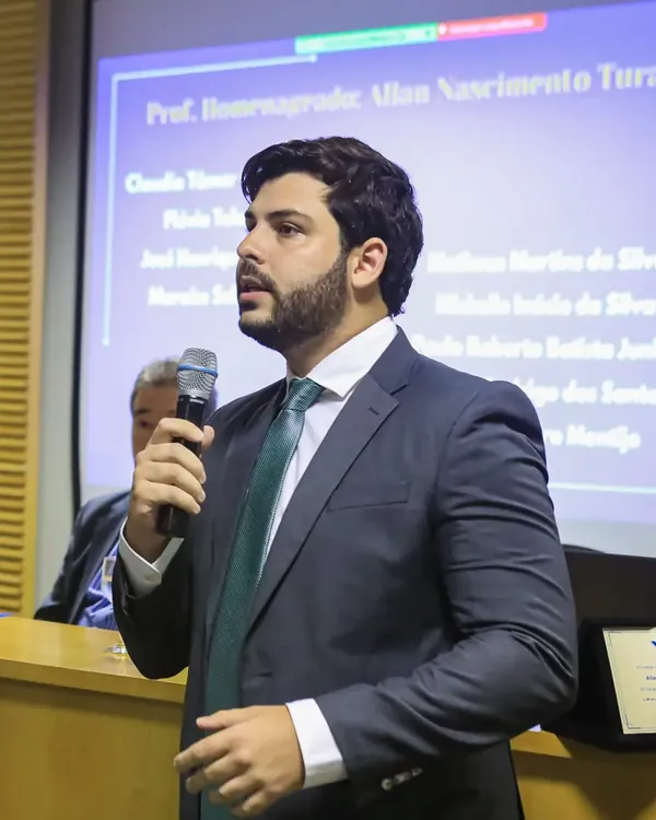 Allan Turano discursa ao microfone diante de slide que o apresenta como professor homenageado