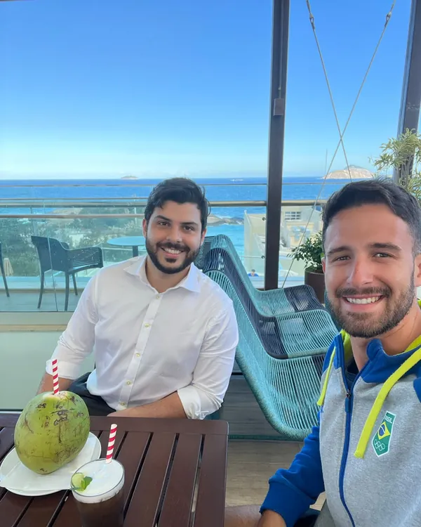 Allan Turano e um convidado com agasalho do Time Brasil, num terraço de frente para o mar