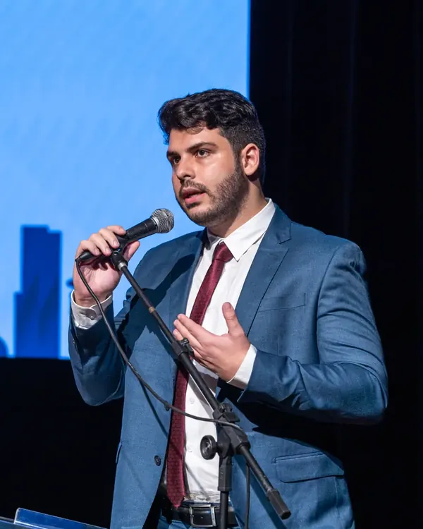 Allan Turano fala ao microfone em um palco, durante palestra